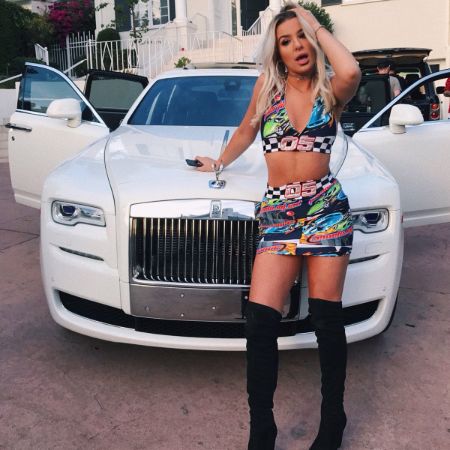 tana mongeau in a bra halter top matching skirt and thigh high boots standing infront of a car 